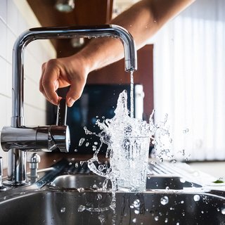 Eine Hand dreht den Wasserhahn auf, wodurch Wasser in ein Glas fließt und dabei spritzende Tropfen erzeugt.