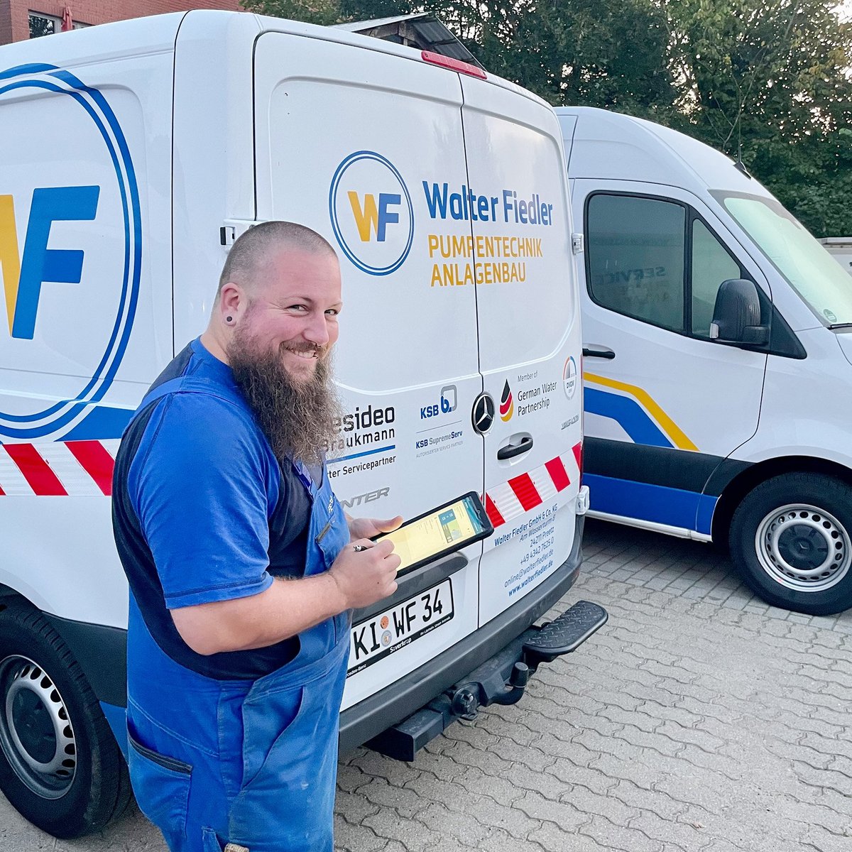 Der Mann in blauer Arbeitskleidung lächelt, während er an einem Lieferwagen mit dem Logo 'Walter Fiedler' steht.