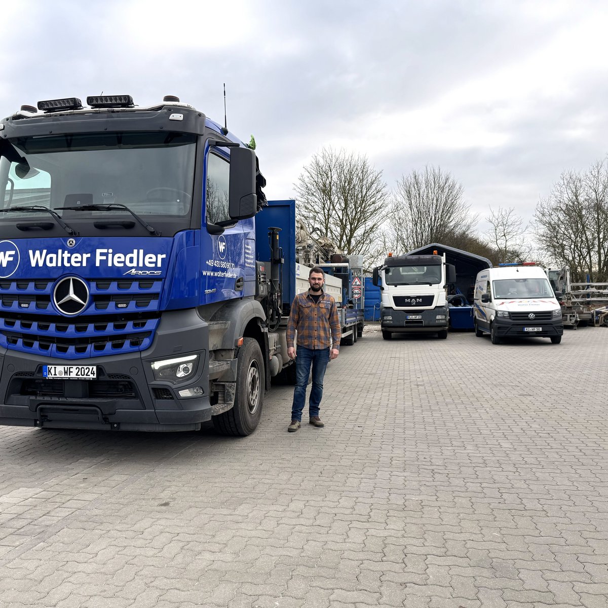 Ein Mann steht auf einem gepflasterten Platz neben einem großen blauen Lkw mit der Aufschrift 'Walter Fiedler'.