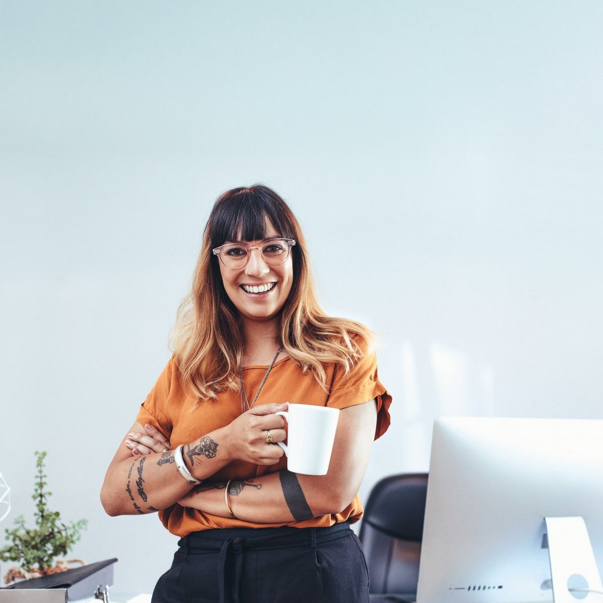 Eine lächelnde Frau mit langen, welligen Haaren steht in einem modernen Büro und hält eine Tasse in der Hand.