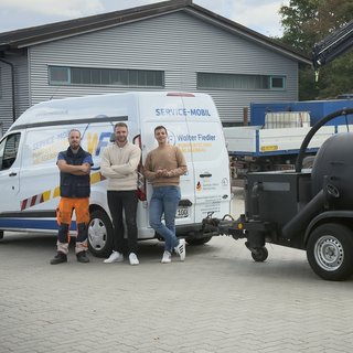 Die Szene zeigt drei Fachleute in Arbeitskleidung, die stolz vor ihrem Service-Mobil und einem speziellen Anhänger stehen.