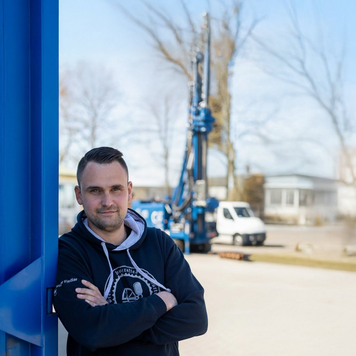 Ein Mann mit einem selbstbewussten Ausdruck lehnt an einer blauen Wand, während ein Baugerät im Hintergrund sichtbar ist.