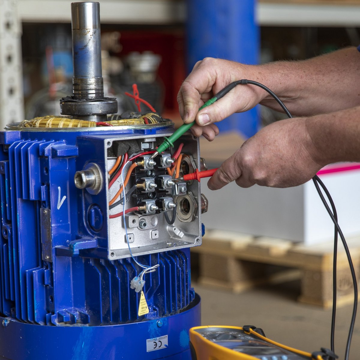 Ein Handwerker überprüft die elektrischen Verbindungen eines blauen Motors, während er ein Messgerät in der Hand hält.